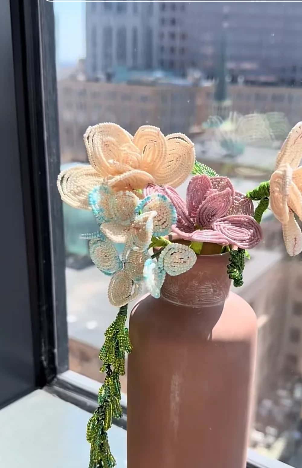 Beaded peach and mauve flowers with trailing green beaded stems in a clay vase, backlit by a city window