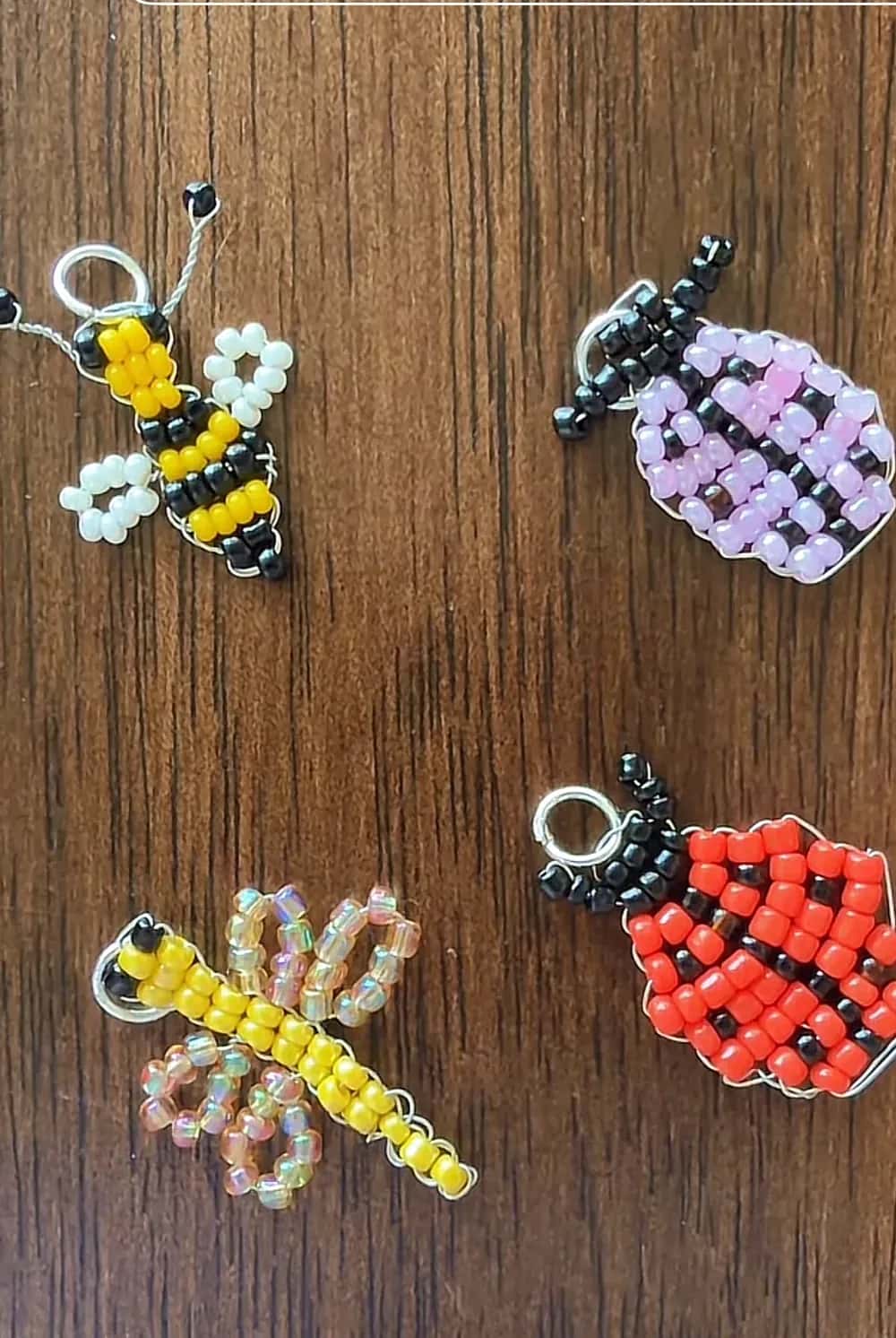 Five beaded bug charms on dark wood: a yellow and black bee, a purple ladybug, a pale dragonfly, a black beetle and a red ladybug
