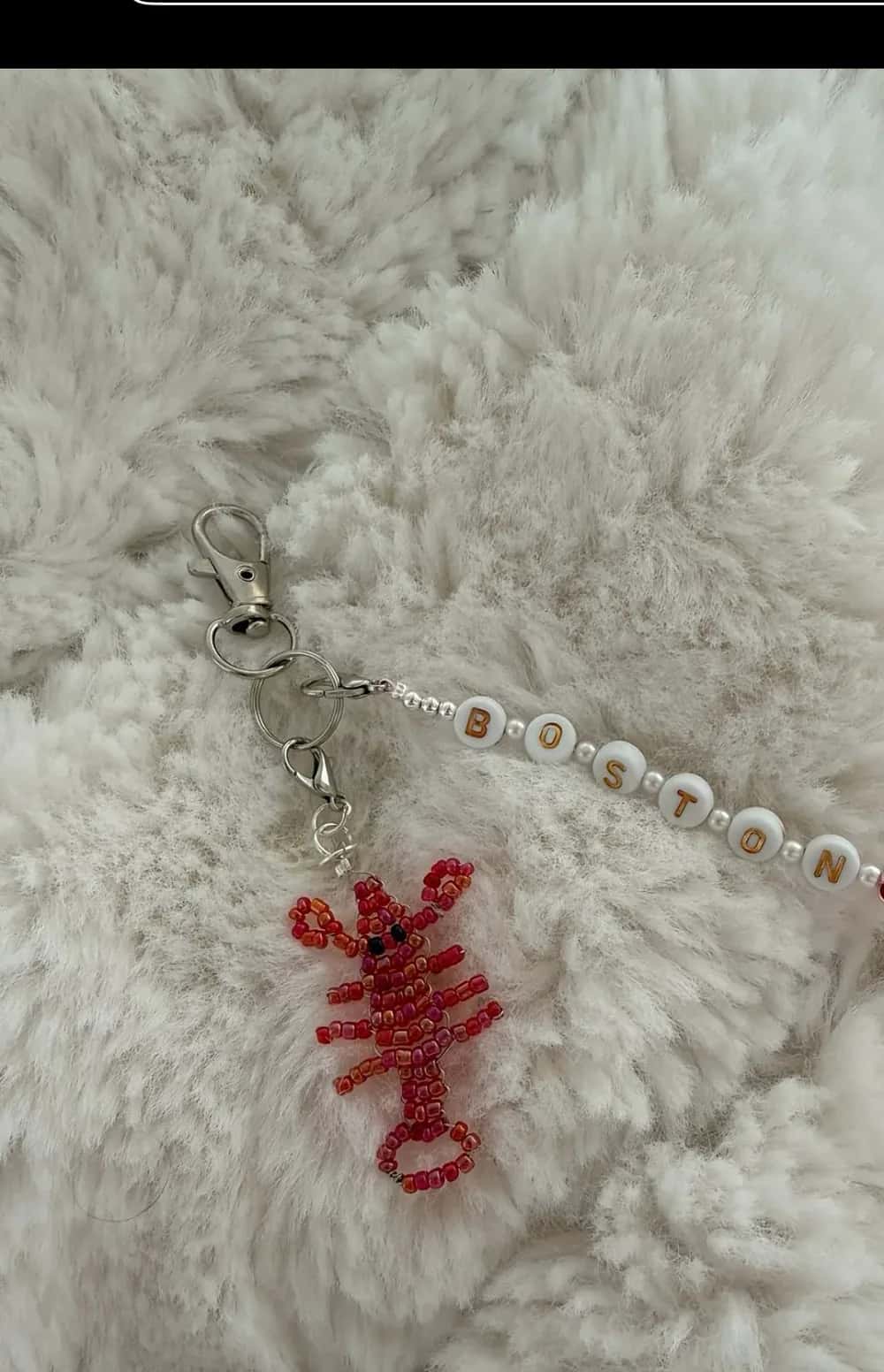 A red beaded lobster charm beside white letter beads spelling BOSTON, on a cream sheepskin background
