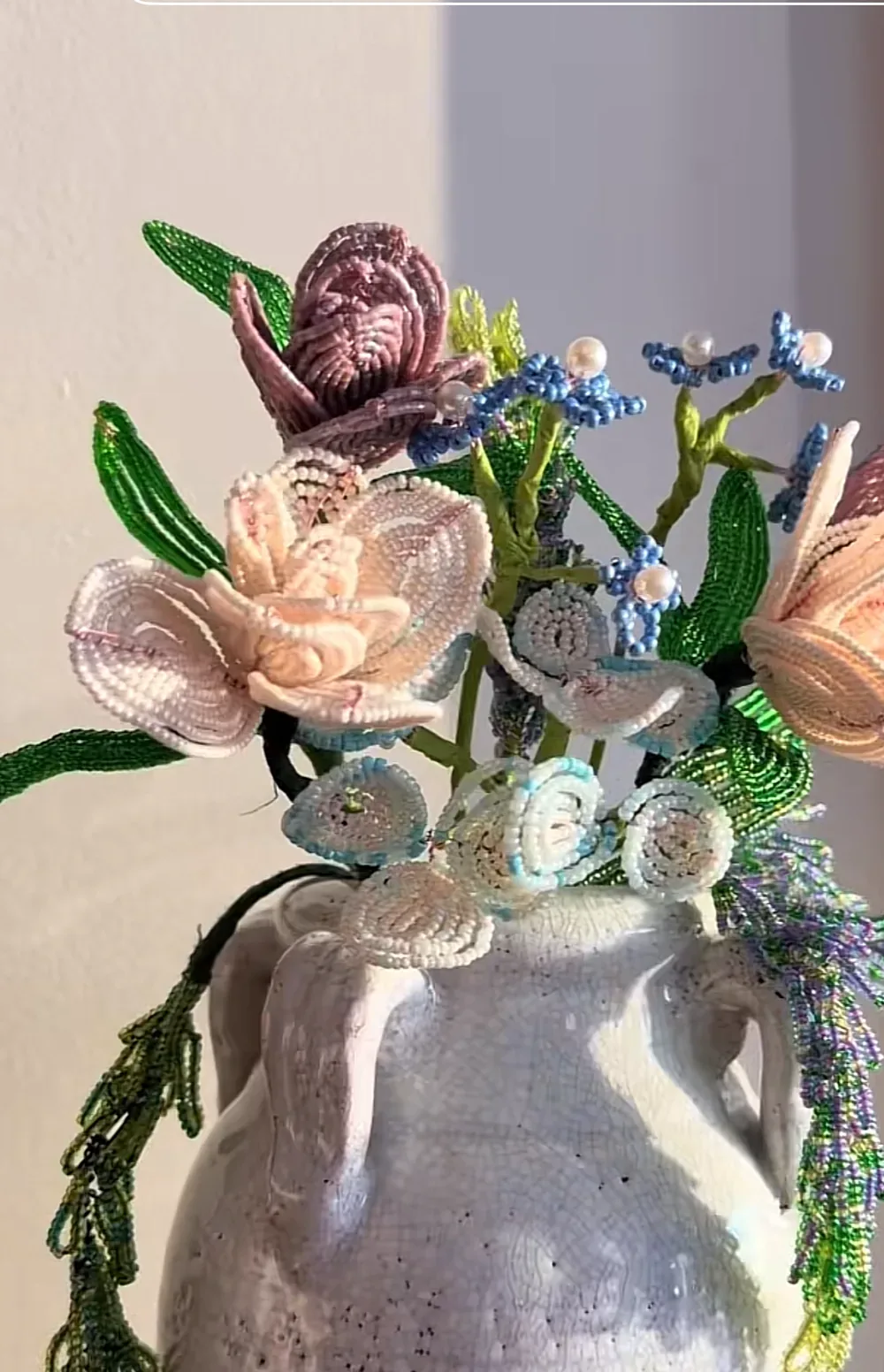 A beaded bouquet of cream and mauve roses with blue muscari and green foliage, arranged in a white stoneware pitcher